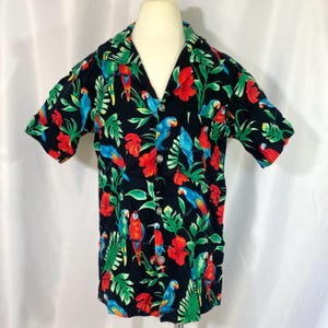 May include: A short-sleeved button-down shirt with a black background, featuring a vibrant tropical print. The design includes colorful parrots, red hibiscus flowers, and green foliage. The shirt is displayed on a mannequin.