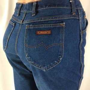 Op de afbeelding: Een paar blauwe denim jeans met een bruin leren patch met daarop "Wrangler" op de achterzak.