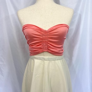 May include: A coral-colored tube top with a sweetheart neckline and gathered detailing. The top is paired with a pair of cream-colored high-waisted pants. The clothing is displayed on a mannequin.