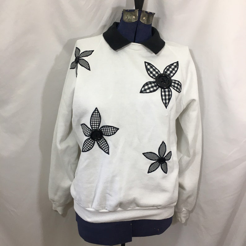 Puede incluir: Sudadera blanca con cuello negro y aplicaciones de flores a cuadros en blanco y negro. Las flores tienen botones negros en el centro.