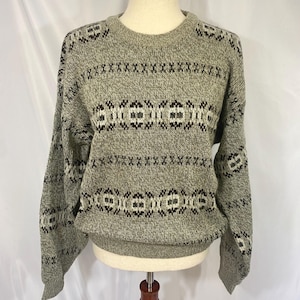 May include: A long-sleeved crew neck sweater in a heathered sage green knit. The garment features horizontal bands of geometric patterns in dark brown, black, and white across the chest, shoulders, and sleeves, with a ribbed collar and cuffs.