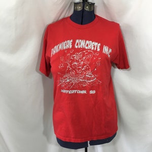May include: Red t-shirt with a white graphic of a muscular figure breaking through concrete. The text "Premiere Concrete Inc. Watertown, SD" is printed on the shirt.