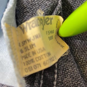 Może przedstawiać: Ż&oacute;łta metka dżins&oacute;w Wrangler z tekstem "13MW JMF F540 008 6 SLIM MADE IN USA 100% COTTON 051071 607025" i "CARE ON REVERSE".