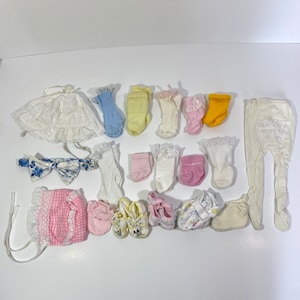 May include: A collection of baby clothes and accessories, including a white dress with lace trim, a pink and white gingham dress, a pair of white tights, a headband with a blue bow, and several pairs of socks and shoes.