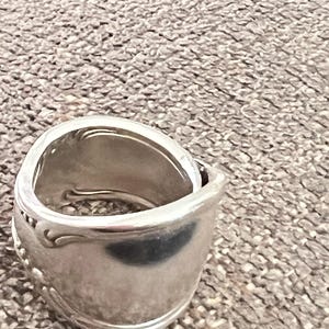 May include: A silver ring crafted from a spoon, featuring a curved design and decorative details. The ring has a polished finish and a unique, handcrafted appearance. The ring is a piece of jewelry.