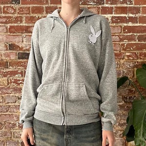 Peut inclure: Un sweat à capuche gris zippé avec un logo de lapin blanc sur la poitrine. Le sweat à capuche a une capuche à cordon et deux poches avant. La personne porte un jean bleu.