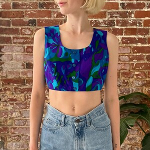 May include: A sleeveless crop top with a colourful abstract floral print in shades of blue, green, and purple. The top has a button closure at the front.