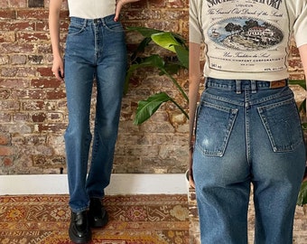 May include: Two women wearing high-waisted blue denim jeans. The woman on the left is wearing a white short-sleeved shirt with a button-up collar. The woman on the right is wearing a white short-sleeved t-shirt with a graphic print of a Southern Comfort liquor bottle label. The label reads "Southern Comfort Liqueur The Grand Old Drink of the South 341 ml Une Tradition du Sud Southern Comfort Corporation 40% Alc./Vol."