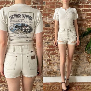 May include: White t-shirt with a vintage Southern Comfort liqueur logo on the back. The logo features a drawing of a house and trees with the text "Southern Comfort Liqueur The Grand Old Drink of the South" and "Une Tradition de Luxe" in a circle around the drawing. The t-shirt is paired with white denim shorts with a distressed look and frayed edges.