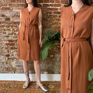 May include: A rust-coloured sleeveless dress with a button-up front and a matching belt. The dress has a v-neck and a midi length.