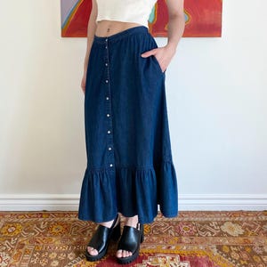May include: A white tank top paired with a dark blue denim maxi skirt. The skirt features a button-down front and a ruffled hem. The model is wearing black platform sandals. The background includes a colorful abstract painting.