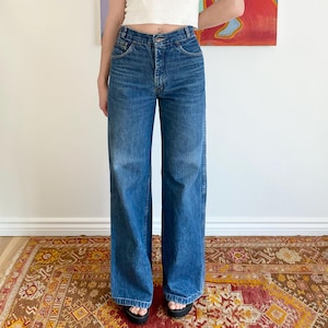 May include: A white ribbed tank top paired with high-waisted, wide-leg blue jeans. The jeans have a classic five-pocket design. The model is wearing black sandals. The background features a colorful abstract painting.