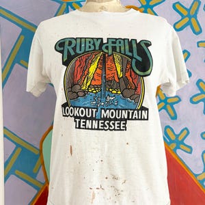 Peut inclure: Un t-shirt blanc à manches courtes avec un motif représentant Ruby Falls à Lookout Mountain, Tennessee. Le design inclut une cascade, des formations rocheuses et le texte "RUBY FALLS" et "LOOKOUT MOUNTAIN TENNESSEE". Le t-shirt présente des taches brunes.