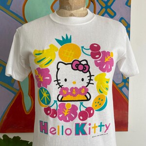 Puede incluir: Camiseta blanca con un dise&ntilde;o de Hello Kitty rodeada de ilustraciones coloridas de frutas y flores. El texto "Hello Kitty" est&aacute; en rosa y turquesa. La camiseta es de cuello redondo.