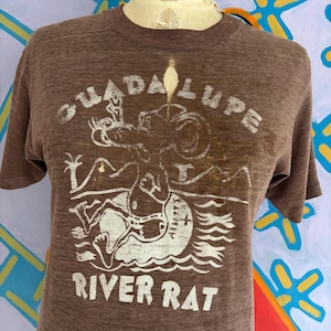 May include: Brown t-shirt with the text "GUADALUPE RIVER RAT" in white. The graphic depicts a rat-like creature in water. The shirt is displayed on a mannequin.