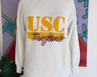 May include: A white sweatshirt with the text "USC Trojans" in yellow and purple. The sweatshirt is displayed on a mannequin. The background features a colorful abstract painting.