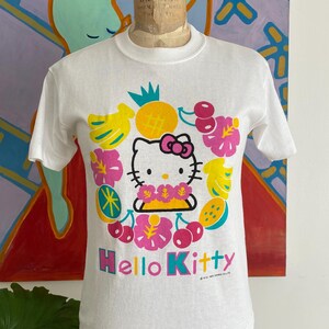Puede incluir: Camiseta blanca con un gr&aacute;fico de Hello Kitty rodeado de frutas y flores tropicales. El dise&ntilde;o incluye pl&aacute;tanos, cerezas y una pi&ntilde;a. Las palabras "Hello Kitty" est&aacute;n impresas en una fuente vibrante y multicolor.