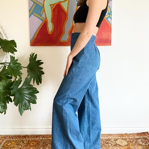 Puede incluir: Pantalones vaqueros azules de cintura alta y pernera ancha. Los pantalones se combinan con un top negro. La modelo est&aacute; de pie frente a una pintura abstracta colorida y una alfombra estampada.