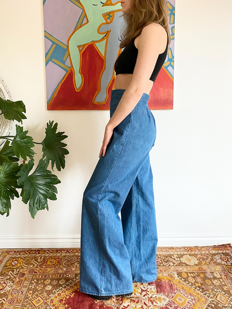Puede incluir: Pantalones vaqueros azules de cintura alta y pernera ancha. Los pantalones se combinan con un top negro. La modelo est&aacute; de pie frente a una pintura abstracta colorida y una alfombra estampada.