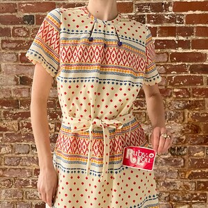May include: A white dress with a geometric pattern in red, orange, blue, and yellow. The dress has a tie at the waist and short sleeves. The dress is made of a lightweight fabric and has a relaxed fit. The dress is perfect for a summer day or a casual event.