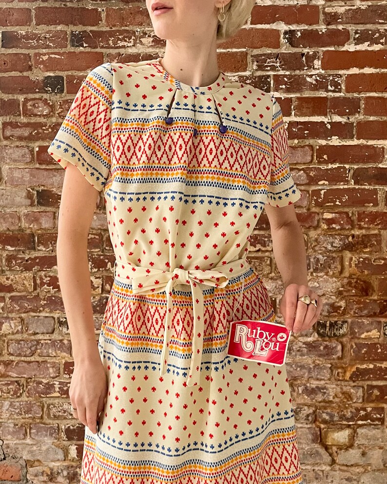May include: A white dress with a geometric pattern in red, orange, blue, and yellow. The dress has a tie at the waist and short sleeves. The dress is made of a lightweight fabric and has a relaxed fit. The dress is perfect for a summer day or a casual event.