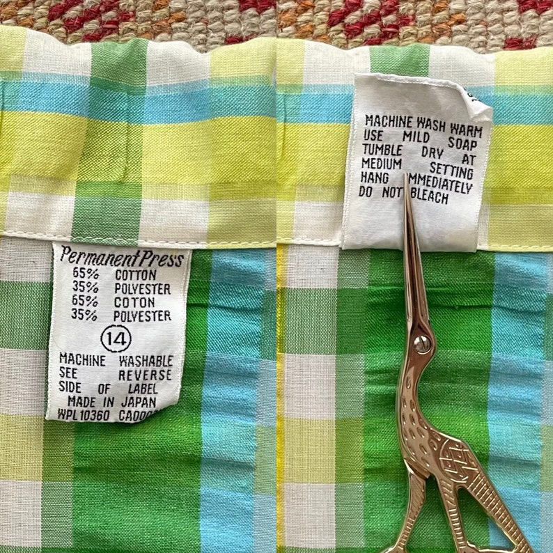 May include: A green, yellow and blue plaid fabric swatch with a white care label that reads "Machine Wash Warm Use Mild Soap Tumble Dry at Medium Setting Hang Immediately Do Not Bleach". A white label with the text "Permanent Press 65% Cotton 35% Polyester 65% Coton 35% Polyester (14) Machine Washable See Reverse Side of Label Made in Japan WPL 10360 CA000" is also visible.