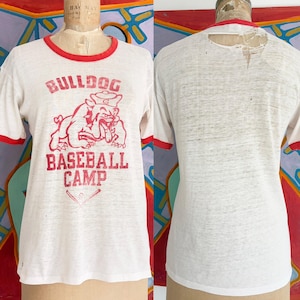 May include: White t-shirt with red trim. The front displays a red bulldog graphic and the text "BULLDOG BASEBALL CAMP". The shirt has a crew neck and short sleeves. The back of the shirt has a small hole near the neckline.