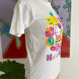 Puede incluir: Camiseta blanca con un gr&aacute;fico de Hello Kitty rodeada de flores y frutas tropicales de colores vibrantes. El texto "Hello Kitty" est&aacute; en rosa y azul. La camiseta se muestra en un maniqu&iacute;.