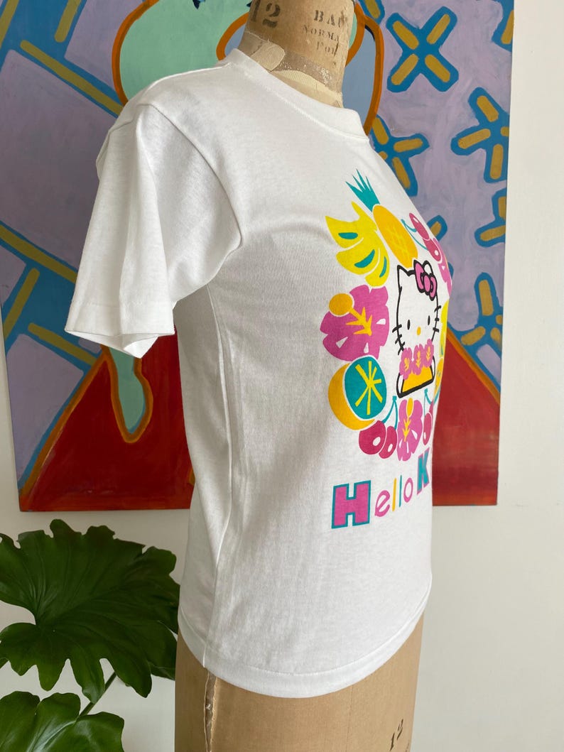 Puede incluir: Camiseta blanca con un gr&aacute;fico de Hello Kitty rodeada de flores y frutas tropicales de colores vibrantes. El texto "Hello Kitty" est&aacute; en rosa y azul. La camiseta se muestra en un maniqu&iacute;.