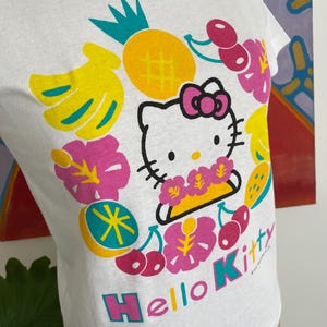 Puede incluir: Camiseta blanca con un dise&ntilde;o de Hello Kitty rodeado de frutas y flores tropicales. El dise&ntilde;o incluye una pi&ntilde;a amarilla, pl&aacute;tanos, cerezas y las palabras "Hello Kitty" en un arco&iacute;ris.