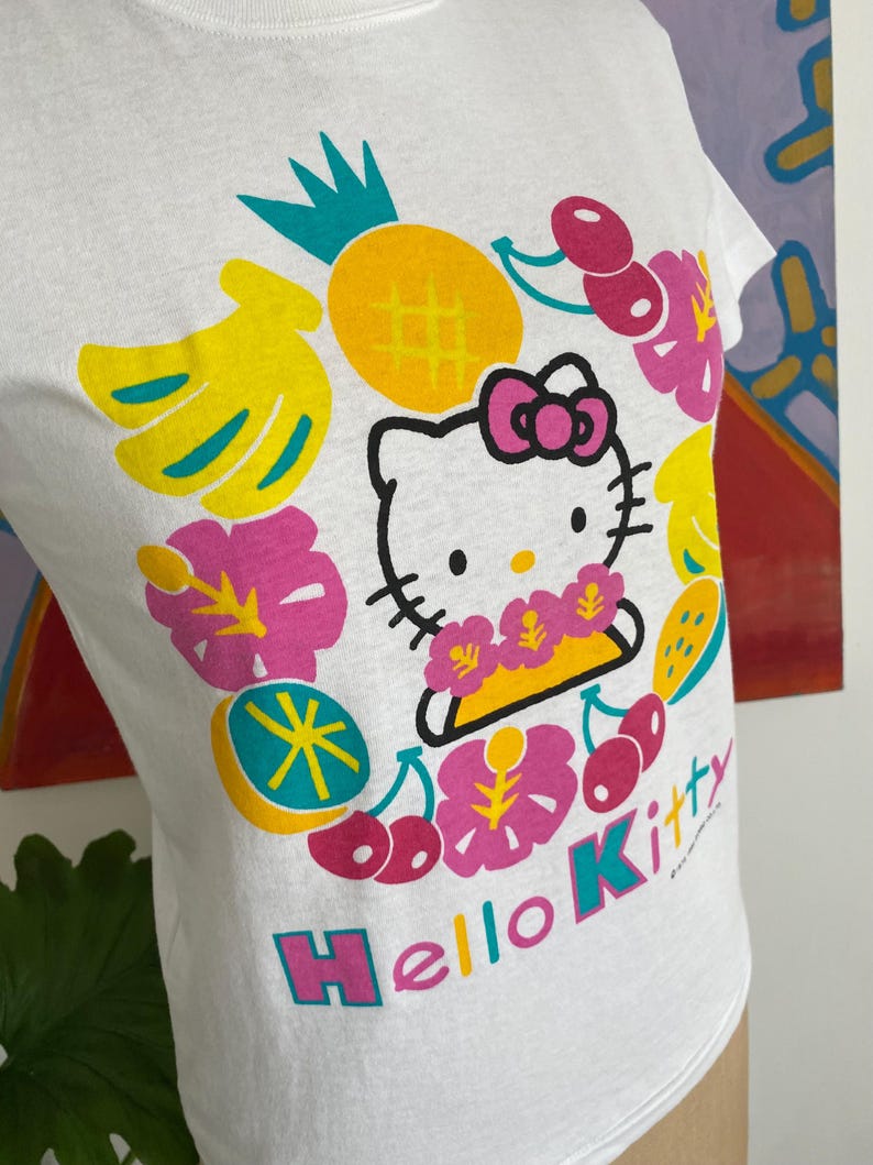 Puede incluir: Camiseta blanca con un dise&ntilde;o de Hello Kitty rodeado de frutas y flores tropicales. El dise&ntilde;o incluye una pi&ntilde;a amarilla, pl&aacute;tanos, cerezas y las palabras "Hello Kitty" en un arco&iacute;ris.