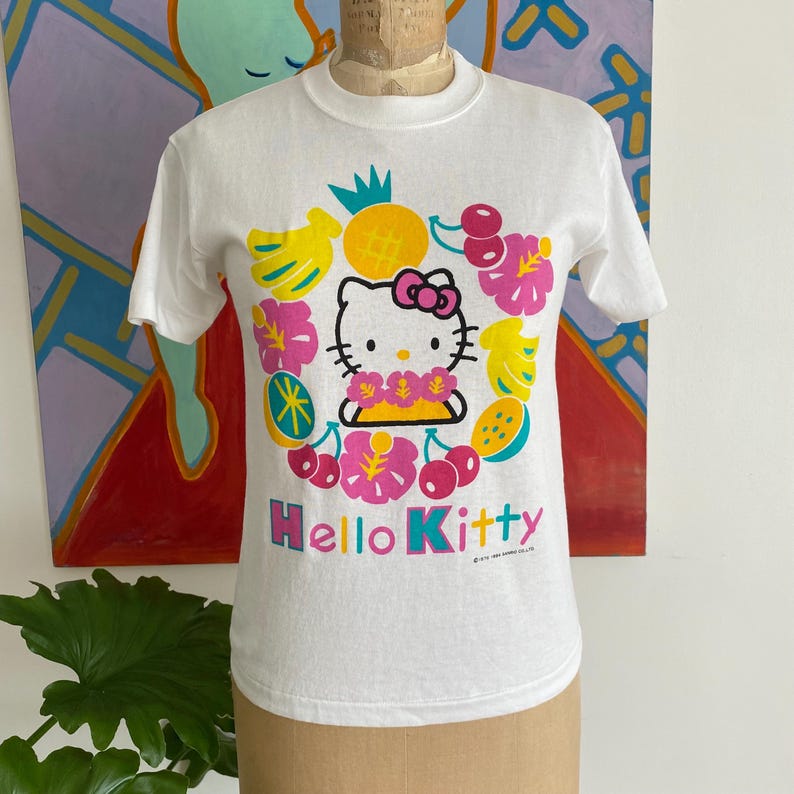 Puede incluir: Camiseta blanca con un gr&aacute;fico de Hello Kitty rodeado de frutas y flores tropicales. El dise&ntilde;o incluye pl&aacute;tanos, cerezas y una pi&ntilde;a. Las palabras "Hello Kitty" est&aacute;n impresas en una fuente vibrante y multicolor.
