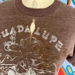 May include: Brown t-shirt with a crew neck and short sleeves. The shirt features a distressed white graphic with the word "GUADALUPE" and a drawing of a figure. The shirt is worn on a mannequin.