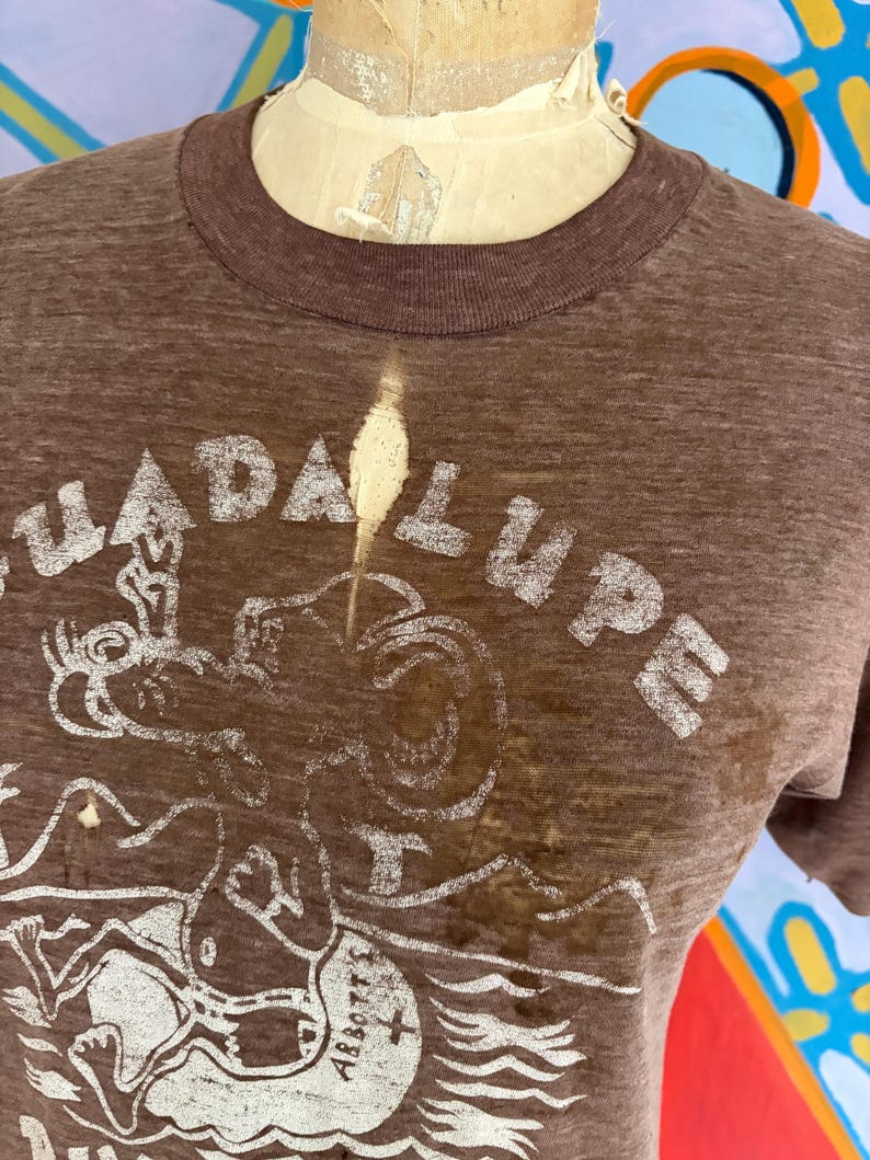 May include: Brown t-shirt with a crew neck and short sleeves. The shirt features a distressed white graphic with the word "GUADALUPE" and a drawing of a figure. The shirt is worn on a mannequin.