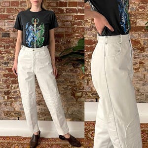 Può includere: Pantaloni in pelle bianca con ginocchiere e una cucitura laterale. I pantaloni sono indossati da una persona che indossa una maglietta nera con una grafica di una spada e due draghi.