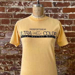 May include: A yellow short-sleeved t-shirt with a black graphic print that reads "OASISPORTSWEAR ULTRA COLOR Thermal Nuclear Science".