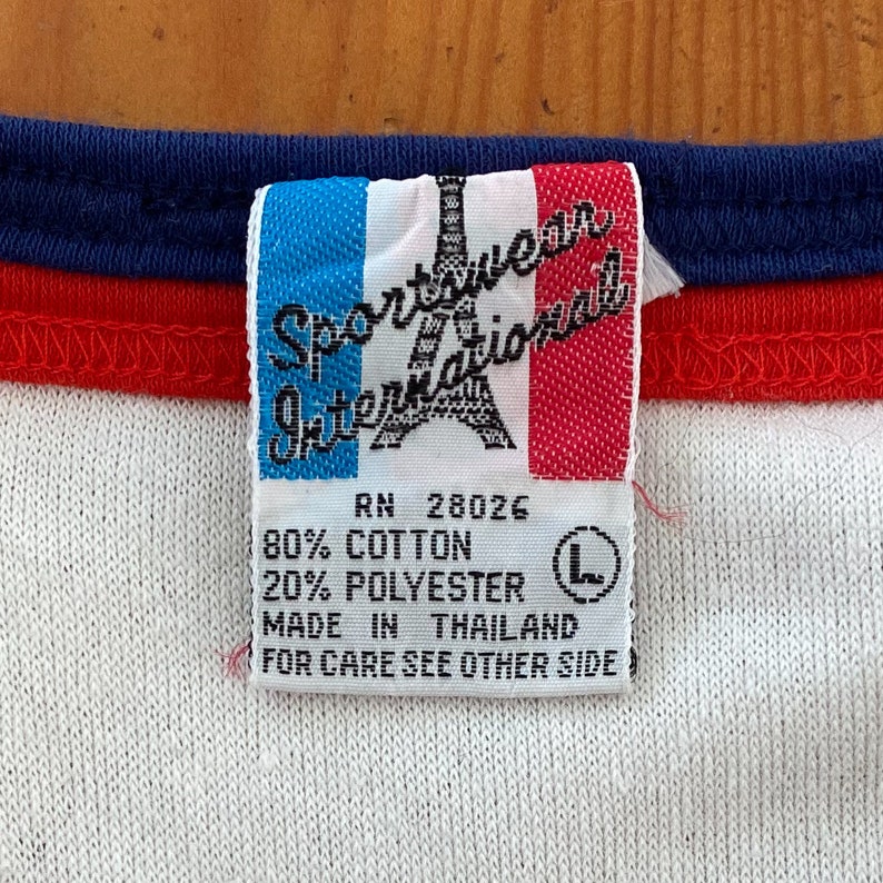 Pu&ograve; includere: Un'etichetta bianca con bordo blu e rosso, che presenta il testo "Sportwear International" con una grafica stilizzata della Torre Eiffel. L'etichetta include anche il testo "RN 28026", "80% Cotone", "20% Poliestere", "Prodotto in Thailandia", e "Per la cura, vedere l'altro lato".