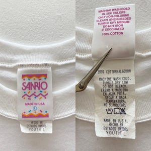 Puede incluir: Primer plano de una camiseta blanca con dos etiquetas adjuntas. Una etiqueta presenta el logotipo de Sanrio en rosa y azul, con "Made in USA" y la talla "L". La otra etiqueta detalla las instrucciones de lavado y "100% Algod&oacute;n".