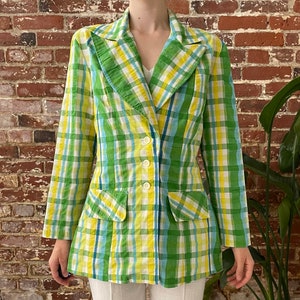 May include: A green and yellow plaid blazer with a white button fastening. The blazer has a collar and two pockets.
