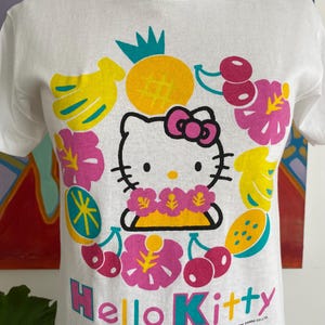 Puede incluir: Camiseta blanca con un gr&aacute;fico de Hello Kitty rodeada de frutas y flores tropicales. El dise&ntilde;o incluye una pi&ntilde;a, pl&aacute;tanos, cerezas y el texto "Hello Kitty". La camiseta es de cuello redondo.