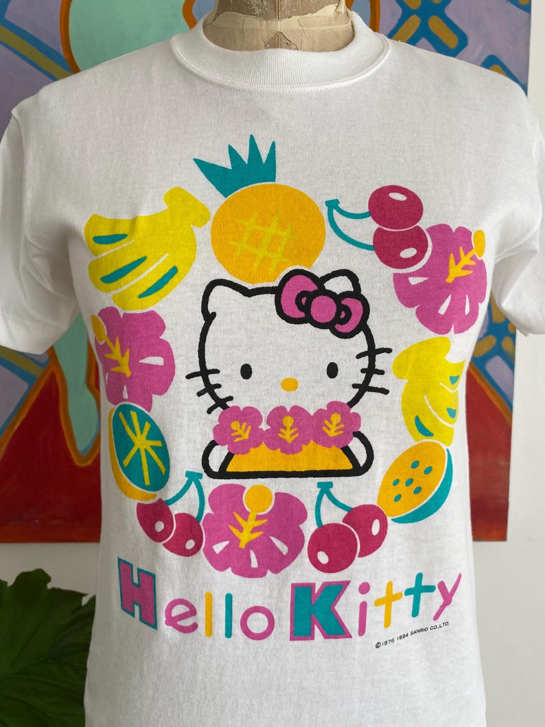Puede incluir: Camiseta blanca con un gr&aacute;fico de Hello Kitty rodeada de frutas y flores tropicales. El dise&ntilde;o incluye una pi&ntilde;a, pl&aacute;tanos, cerezas y el texto "Hello Kitty". La camiseta es de cuello redondo.