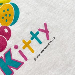 Puede incluir: Camiseta blanca con un gr&aacute;fico colorido de frutas y la palabra "Kitty" en rosa, amarillo y turquesa. El texto "&copy;1976 1994 SANRIO CO.,LTD." est&aacute; impreso en negro. La camiseta tiene una est&eacute;tica vintage.