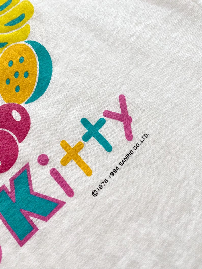 Puede incluir: Camiseta blanca con un gr&aacute;fico colorido de frutas y la palabra "Kitty" en rosa, amarillo y turquesa. El texto "&copy;1976 1994 SANRIO CO.,LTD." est&aacute; impreso en negro. La camiseta tiene una est&eacute;tica vintage.