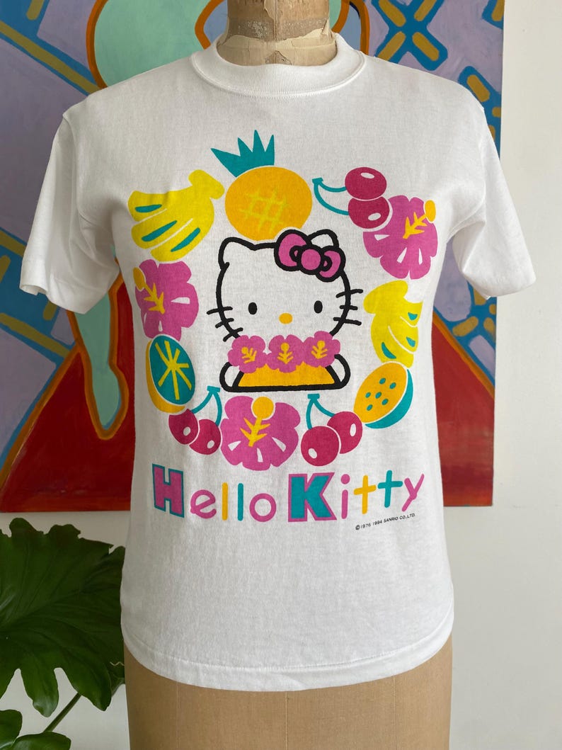 Puede incluir: Camiseta blanca con un dise&ntilde;o de Hello Kitty rodeada de ilustraciones coloridas de frutas y flores. El texto "Hello Kitty" est&aacute; en rosa y turquesa. La camiseta es de cuello redondo.