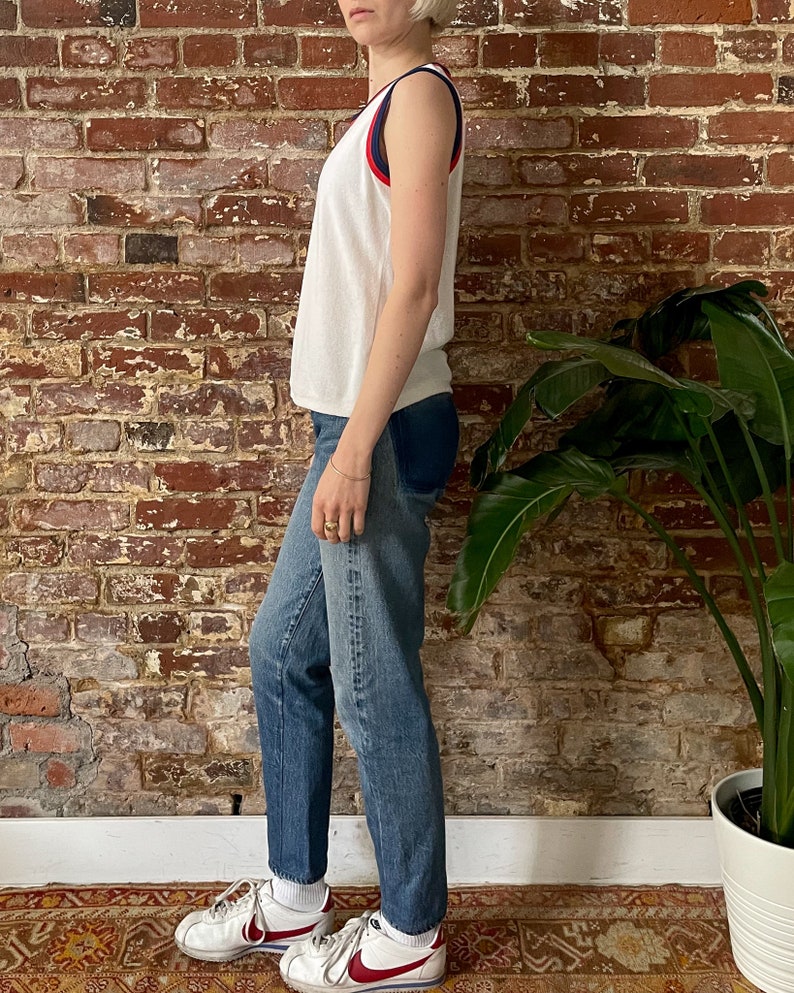Pu&ograve; includere: Canotta bianca senza maniche con bordini rossi e blu. La canotta &egrave; indossata con jeans blu e scarpe da ginnastica bianche.