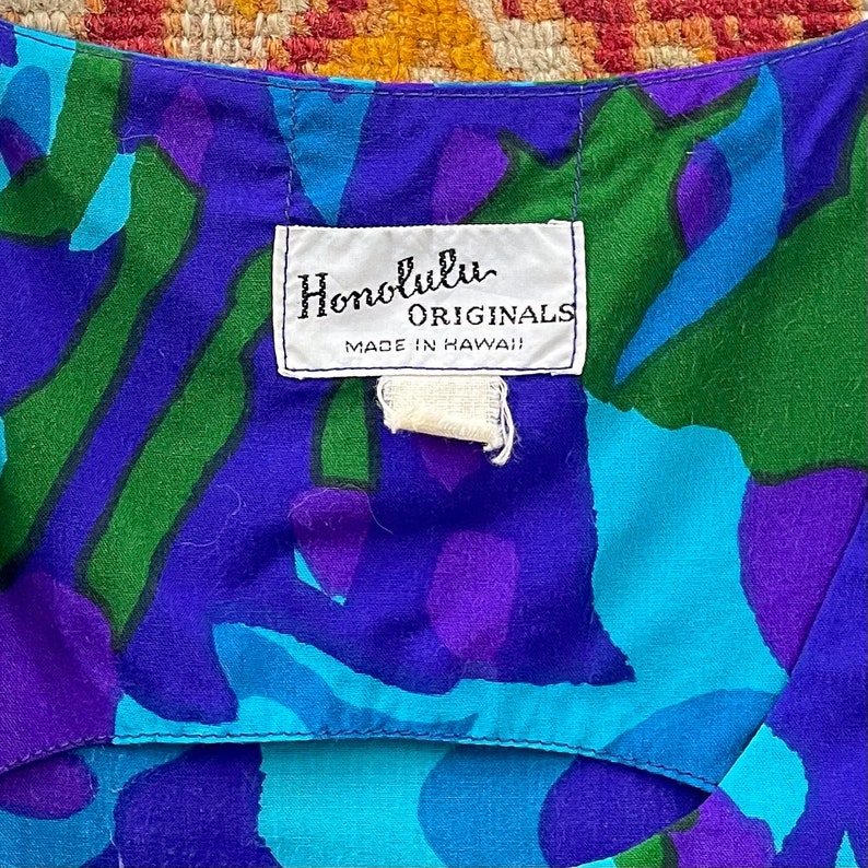Peut inclure: Un tissu &agrave; motifs bleus, verts et violets avec une &eacute;tiquette blanche qui indique "Honolulu Originals Made in Hawaii!"