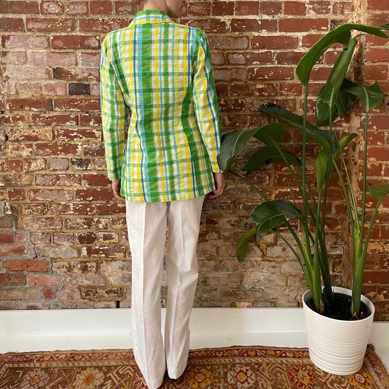 May include: A green, yellow and blue plaid blazer with long sleeves and a white button-up collar. The blazer is worn over a pair of white trousers.