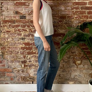 Pu&ograve; includere: Canotta bianca senza maniche con bordini rossi e blu. La canotta &egrave; realizzata in un tessuto morbido e testurizzato. La modella indossa jeans blu e scarpe da ginnastica bianche.