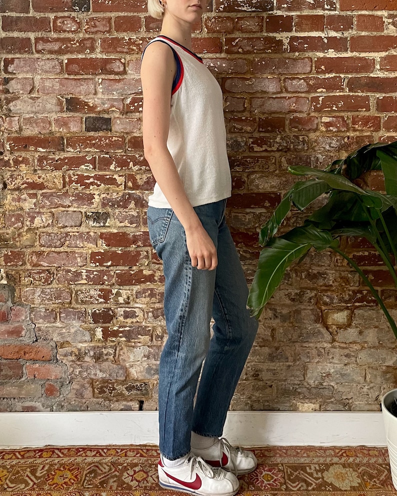 Pu&ograve; includere: Canotta bianca senza maniche con bordini rossi e blu. La canotta &egrave; realizzata in un tessuto morbido e testurizzato. La modella indossa jeans blu e scarpe da ginnastica bianche.