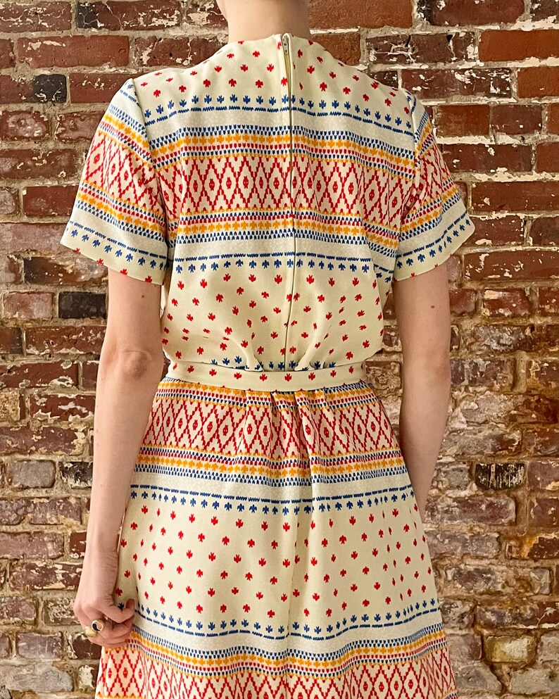 May include: A white dress with a geometric pattern in red, blue, and yellow. The dress has a short sleeve and a fitted bodice. The skirt is full and flows to the knees.